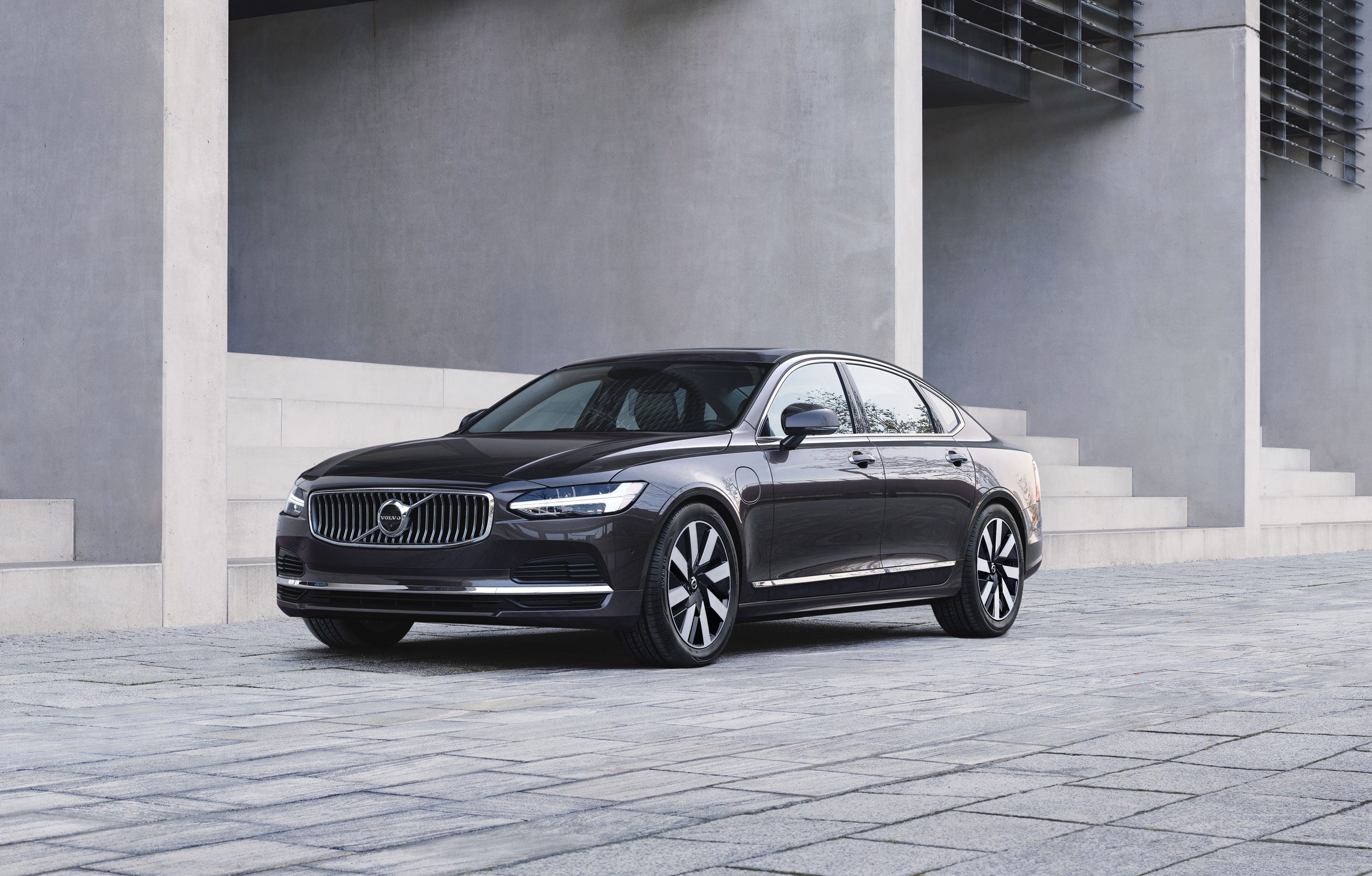 Un sedan Volvo S90 Recharge T8 AWD gri este parcat în fața unei clădiri din beton.